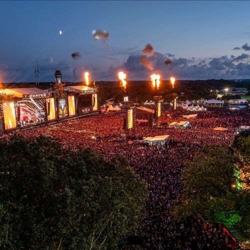 Aerial view of Wacken Open Air 2025 at dusk, showing a massive crowd, brightly lit stages, and pyrotechnic flames shooting into the sky.