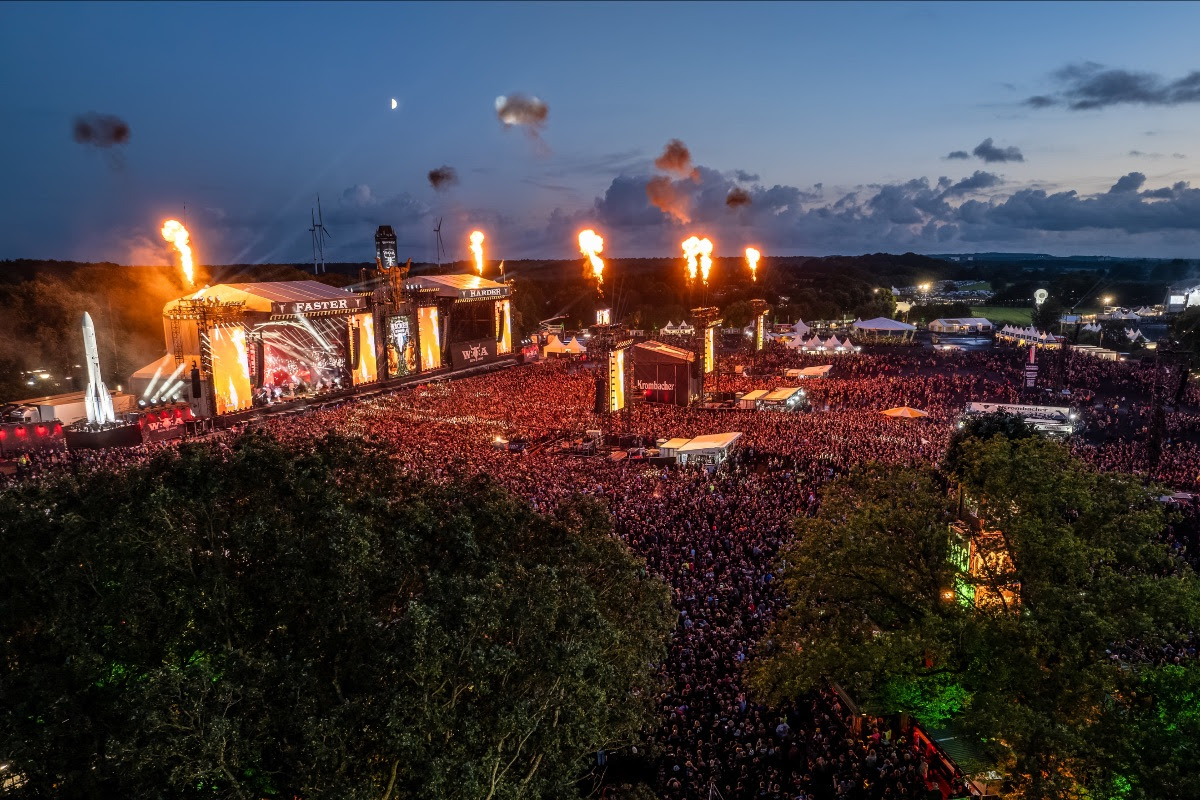 Aerial view of Wacken Open Air 2025 at dusk, showing a massive crowd, brightly lit stages, and pyrotechnic flames shooting into the sky.